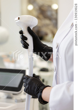 Close-up cosmetologist in white coat and black gloves holding modern laser handpiece device for beauty treatments, professional skincare technology concept in aesthetic medicine and dermatology clinic Close-up cosmetologist in white coat and black gloves holding modern laser handpiece device for beauty treatments, professional skincare technology concept in aesthetic medicine and dermatology clinic 130919828