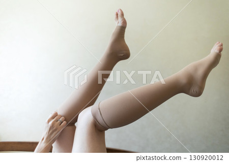 Woman putting on compression stockings in bedroom to prevent varicose veins. Swelling and leg pain, medical hosiery for circulation and vein health 130920012