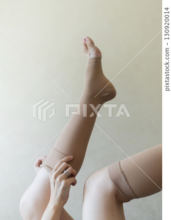 Woman putting on compression stockings in bedroom to prevent varicose veins. Swelling and leg pain, medical hosiery for circulation and vein health Woman putting on compression stockings in bedroom to prevent varicose veins. Swelling and leg pain, medical hosiery for circulation and vein health 130920014
