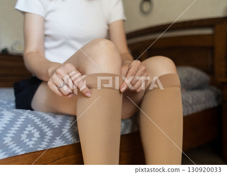 Woman putting on compression stockings in bedroom to prevent varicose veins. Swelling and leg pain, medical hosiery for circulation and vein health 130920033