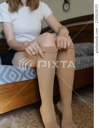 Woman putting on compression stockings in bedroom to prevent varicose veins. Swelling and leg pain, medical hosiery for circulation and vein health Woman putting on compression stockings in bedroom to prevent varicose veins. Swelling and leg pain, medical hosiery for circulation and vein health 130920035