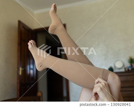 Woman putting on compression stockings in bedroom to prevent varicose veins. Swelling and leg pain, medical hosiery for circulation and vein health Woman putting on compression stockings in bedroom to prevent varicose veins. Swelling and leg pain, medical hosiery for circulation and vein health 130920041