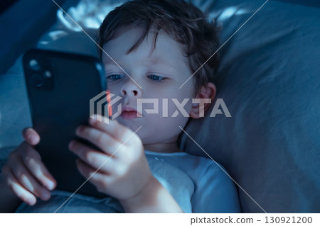 Little boy lying in bed at night, intently watching smartphone screen Little boy lying in bed at night, intently watching smartphone screen 130921200