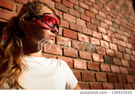 Young woman wearing red sports glasses standing against a brick wall exterior 130922038
