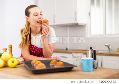 Smiling young woman in apron eating cupcakes from baking tray 130922818