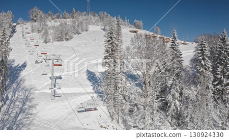 Chairlift transporting skiers uphill on snowy mountain slope, surrounded by snow covered trees and charming chalets, bright sunny day. Bukovel Ukraine ski resort. Winter wild nature travel background 130924330
