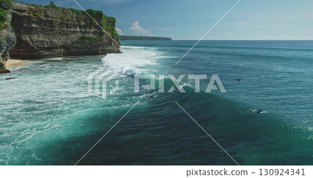 Turquoise ocean waves rolling onto Pantai Dreamland Beach in Bali, with surfers eagerly waiting to ride the waves against a backdrop of cliffs and lush vegetation 130924341