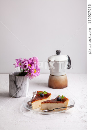 Slices of homemade Basque burnt cheesecake with blueberries and mint leaves, geyser coffee maker, flowers in a vase 130927986