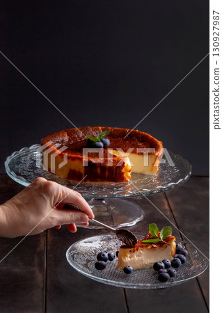 Homemade Basque burnt cheesecake with cream cheese, eggs and cream baked in a crumb crust, with blueberries and mint leaves. Woman breaks off a piece on a plate on dark background 130927987