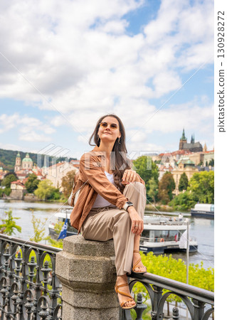 Young woman with a small backpack sitting on a stone railing with Prague Castle in the blurred background. Concept of solo travel, peaceful exploration and discovering historical European cities 130928272