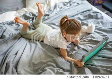 Little girl lying on her parents bed and watching cartoons on a tablet. Concept of screen time, digital entertainment and relaxation during early childhood. 130928281