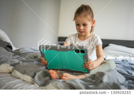Little girl using a tablet while sitting on her parents bed in the bedroom. Concept of screen time, digital play and modern childhood in a home environment. 130928283