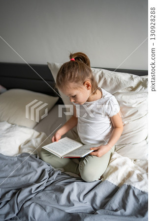 Little girl sitting on her parents bed and reading a book in the bedroom. Concept of quiet time, concentration and love for reading in early childhood. 130928288