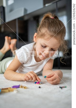 Little girl sitting on the floor and playing with building blocks in the living room. Concept of early development, creativity and focused play in a home environment. Little girl sitting on the floor and playing with building blocks in the living room. Concept of early development, creativity and focused play in a home environment. 130928292