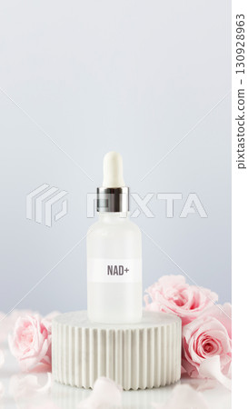 NAD essence in glass droplet bottle surrounded by pink roses on soft grey background. Concept of NAD beauty products, cellular beauty, anti-aging trends, and modern wellness products 130928963