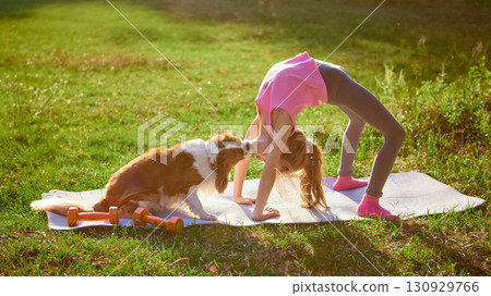 Girl practicing yoga bridge pose on mat with dog kissing her face outdoors 130929766