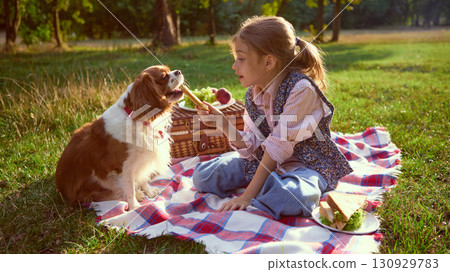 Girl feeding dog on picnic blanket with fruits in summer park Girl feeding dog on picnic blanket with fruits in summer park 130929783