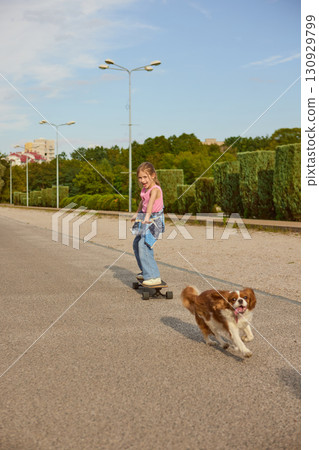 Girl riding skateboard with dog running beside in city park 130929799