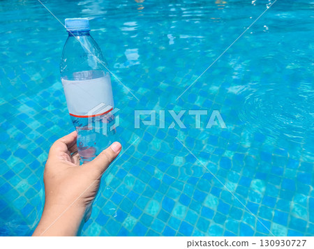 A hand lifts a plastic bottle from clear blue pool water, symbolizing pollution cleanup and environmental awareness. 130930727