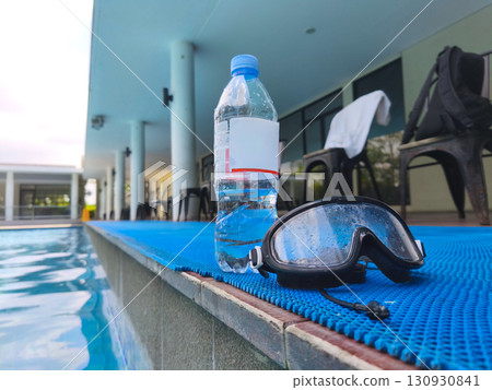 Plastic water bottle and wet swimming goggles on poolside mat, symbolizing summer recreation, hydration, and swimming activities. Plastic water bottle and wet swimming goggles on poolside mat, symbolizing summer recreation, hydration, and swimming activities. 130930841