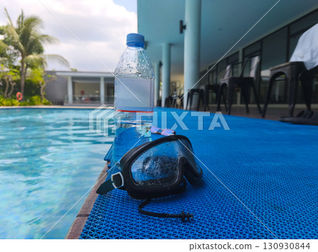 Plastic water bottle and wet swimming goggles on poolside mat, symbolizing summer recreation, hydration, and swimming activities. 130930844
