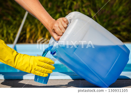 Pouring blue pool chemical from large canister into measuring cup with yellow protective gloves, precise dosage for water treatment, outdoor maintenance near clear swimming pool Pouring blue pool chemical from large canister into measuring cup with yellow protective gloves, precise dosage for water treatment, outdoor maintenance near clear swimming pool 130931105