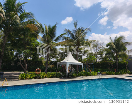 Tropical outdoor swimming pool surrounded by lush palm trees under bright sunny sky, creating a relaxing vacation atmosphere. Tropical outdoor swimming pool surrounded by lush palm trees under bright sunny sky, creating a relaxing vacation atmosphere. 130931142