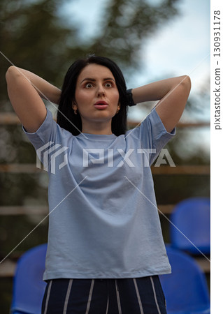 A brunette girl wearing a blue T shirt stands in an empty stadium her hands raised behind her head. She appears shocked and delighted by her unexpected win capturing a moment of excitement and joy. 130931178