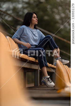 A brunette girl sits on the bleachers of a quiet stadium wearing a blue T shirt and casual pants. She looks thoughtfully into the distance as the sun sets creating a serene atmosphere. 130931179
