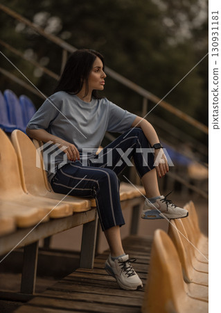 brunette girl relaxes alone in the empty stands of a stadium. She wears a blue t shirt and casual pants enjoying a peaceful moment in the sports venue. brunette girl relaxes alone in the empty stands of a stadium. She wears a blue t shirt and casual pants enjoying a peaceful moment in the sports venue. 130931181