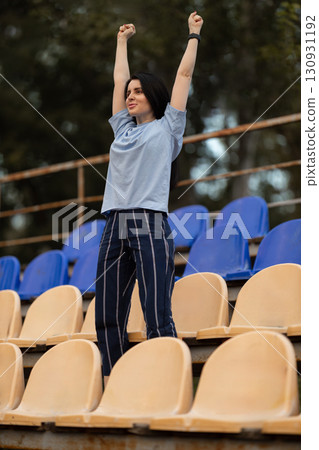 brunette girl wearing blue T shirt stands on empty podium stadium with her hands raised high in celebration expressing pure joy and excitement in moment. brunette girl wearing blue T shirt stands on empty podium stadium with her hands raised high in celebration expressing pure joy and excitement in moment. 130931192