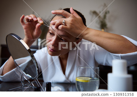 Woman Grooming Eyebrows Using Tweezers in Front of a Mirror at Home Woman Grooming Eyebrows Using Tweezers in Front of a Mirror at Home 130931358