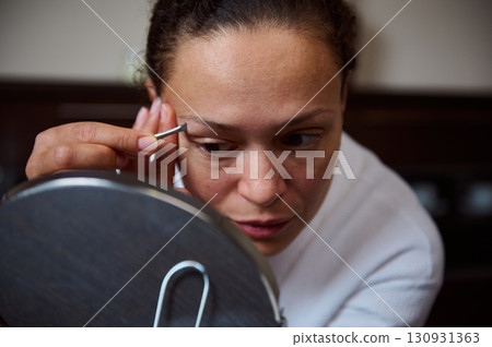 Young Woman Grooming Eyebrows Using Tweezers in Front of a Mirror 130931363