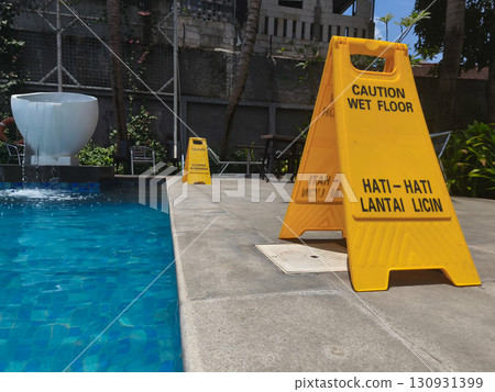 Yellow caution sign reading "Cleaning in Progress" placed near the edge of a swimming pool, emphasizing safety and maintenance in an outdoor area. Yellow caution sign reading "Cleaning in Progress" placed near the edge of a swimming pool, emphasizing safety and maintenance in an outdoor area. 130931399