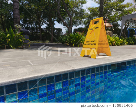 Yellow caution sign reading "Cleaning in Progress" placed near the edge of a swimming pool, emphasizing safety and maintenance in an outdoor area. 130931406