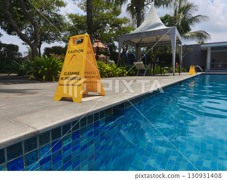 Yellow caution sign reading "Cleaning in Progress" placed near the edge of a swimming pool, emphasizing safety and maintenance in an outdoor area. Yellow caution sign reading "Cleaning in Progress" placed near the edge of a swimming pool, emphasizing safety and maintenance in an outdoor area. 130931408