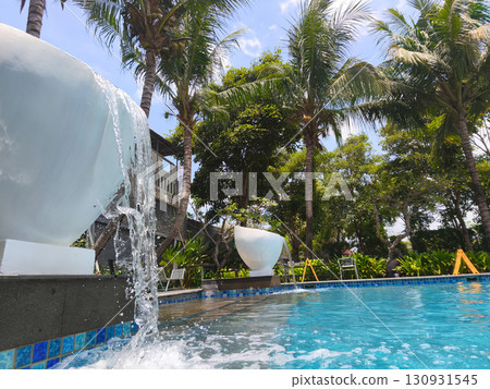 Refreshing water cascading from a fountain into a clear blue swimming pool, surrounded by lush palm trees under a bright sunny sky. 130931545