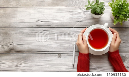 Two hands gently cradle a steaming cup of tea above a wooden table, accompanied by a pen and green plants in the background 130933180