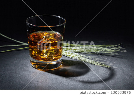 glass of amber whiskey and ears of rye on a black stone table. glass of amber whiskey and ears of rye on a black stone table. 130933976