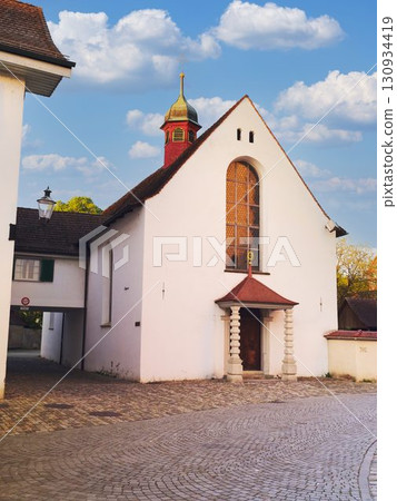 Charming white church with a unique steeple and cobblestone street near a picturesque village in late afternoon light Charming white church with a unique steeple and cobblestone street near a picturesque village in late afternoon light 130934419