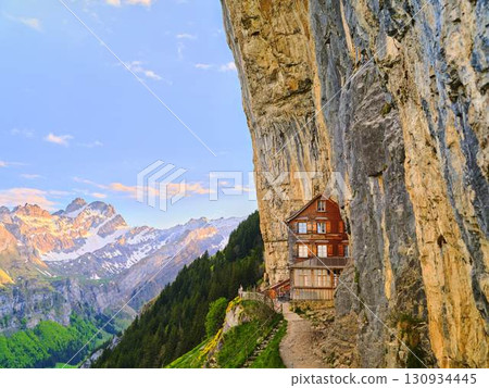 Unique wooden house clings to cliffside with breathtaking mountain views at sunset 130934445