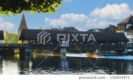 Historic wooden bridge crosses tranquil river under blue sky with fluffy clouds in a quaint village at dawn 130934471