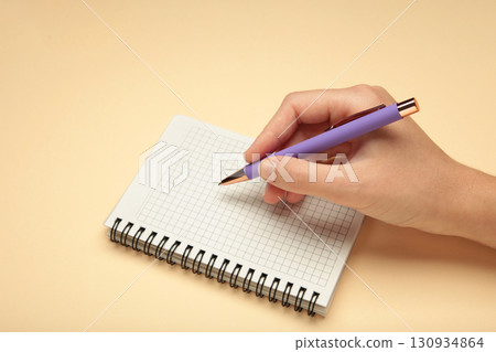Female's hand writing in white blank open spiral notebook with pen on pastel beige background. Flat lay, copy space. Female's hand writing in white blank open spiral notebook with pen on pastel beige background. Flat lay, copy space. 130934864