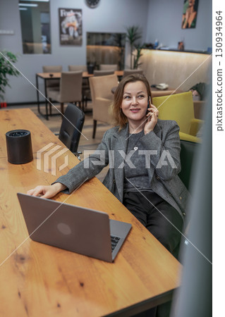 Businesswoman Laptop Phone: Smiling woman talks indoors during daytime while working on project, in office. Businesswoman Laptop Phone: Smiling woman talks indoors during daytime while working on project, in office. 130934964