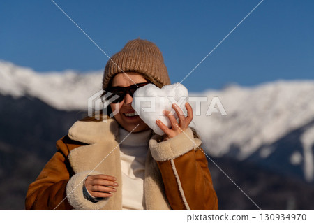 Snow Heart Mountains Winter: Woman playfully holds heart-shaped snowball, sunny day, joyful winter activity. 130934970
