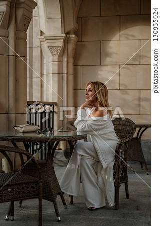 Woman White Outfit Patio Elegant Woman Sitting at a Table in a White Outfit on a Patio with Stone Columns. Woman White Outfit Patio Elegant Woman Sitting at a Table in a White Outfit on a Patio with Stone Columns. 130935024