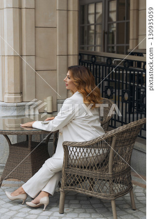 Woman White Suit Outdoor Cafe - Stylish woman in a white suit sitting on a patio chair at an outdoor cafe. Woman White Suit Outdoor Cafe - Stylish woman in a white suit sitting on a patio chair at an outdoor cafe. 130935059