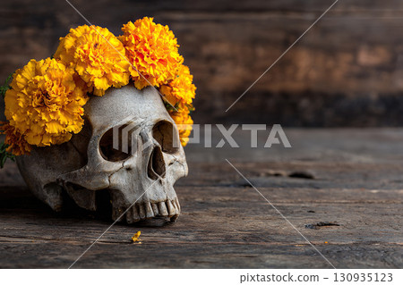A human skull with a wreath of orange flowers stands on a wooden table. Concept for Day of the Dead, Dia De Los Muertos, Diwali, and Halloween decor. A human skull with a wreath of orange flowers stands on a wooden table. Concept for Day of the Dead, Dia De Los Muertos, Diwali, and Halloween decor. 130935123