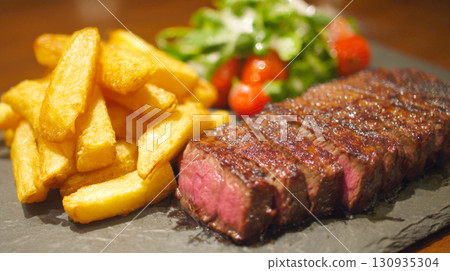 A delicious steak and fries dinner, featuring a perfectly grilled steak cooked to a juicy medium-rare, served alongside crispy golden french fries and a fresh salad, on rustic black plate 130935304