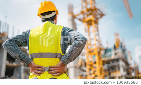 A construction worker, seen from behind, wearing a hard hat and high-visibility vest, holding his lower back in pain while observing a bustling construction site with cranes and buildings 130935660